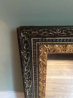 Close-up photo of the upper left corner of an ornately carved mirror frame showing black and bronze floral vine detailing with a beaded strip and inner gold leaf carvings.