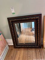 Full shot of the square carved wood framed mirror leaning against a wall on wooden floor.