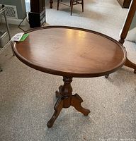 Wood side table oval top from angled view showing latch on top edge and pedestal base with three legs.