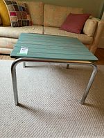 Side view of square side table showing stained wood plank top and chromed tubular metal legs with some pitting.