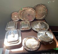 Photo showing multiple silver plate trays, three rectangular serving dishes with lids, large covered serving dish and covered condiment dish arranged on table.