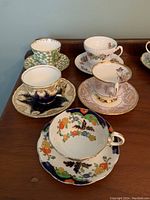 Five fine china teacups and saucers displayed on wooden surface showing variety of floral patterns and colors.