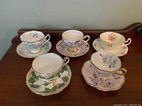 Five fine china teacups and saucers arranged on wooden surface, showing various floral and leaf patterns with gold accents.