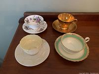 Four fine china teacups with matching saucers on wooden surface; includes floral, gold-tone, and trimmed designs.