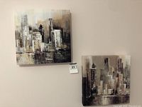 Two small square canvas paintings hung on a wall, showing abstract buildings and cityscape in muted neutral tones.