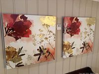 Front view of two matching floral canvas prints with red flowers and gold accents on a beige background, hanging side by side on a white paneled wall.