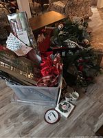 Wide view showing clear plastic bin filled with Christmas wrapping paper, gift bags, and Christmas wreath with ornaments, bows, and pine cones.