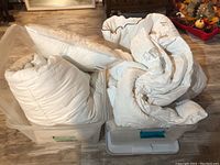 Two clear plastic storage totes containing bundled white duvets and a pillow