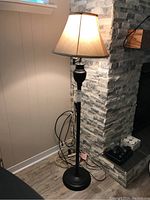 Full view of the black metal floor lamp standing next to a stone wall and wooden floor, showing the beige fabric lampshade lit from inside.