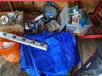 Wide view showing an assortment of hardware and electrical items stored in plastic bins and boxes on a red dolly, blue tarps folded and laid out, lighting fixture, and other small parts such as bulbs and tools.