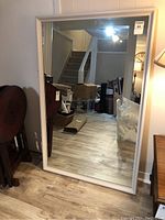 Front view of large white framed mirror standing against a wall reflecting the room