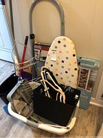 Photo showing small ironing board with polka dot cover, laundry basket filled with iron, fabric box and shoe rack, step ladder in background