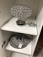 Two light fixtures stored on shelves, one spherical with crystal accents and chrome mount, the other a round crystal panel with mounting and wiring visible.