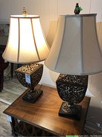 Two matching ornate table lamps with beige fabric bell-shaped shades on a wooden table, photographed in indoor lighting.