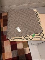 Stacked view of three mats showing the top grayish beige mat and partially visible beige mat with blue pattern beneath, and a corner of the beige and brown geometric mat under them.