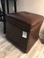 Brown square ottoman with faux leather upholstery, tufted top, nailhead trim, and a label with lot number 34.