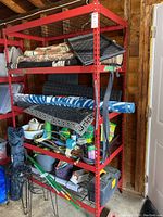 Full view of red five-shelf steel storage unit filled with some rolled mats, containers, and household items.