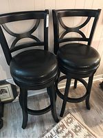 Front view of two black wooden swivel bar stools, showing curved backrests and padded seats.