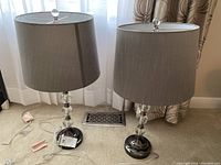 Pair of table lamps standing side by side on carpet, showing the full lamp and grey shade with dark nickel bases and glass ball elements