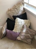 Seven throw pillows piled in corner, showing mix of purples, blacks, and cream with shaggy and woven textures.