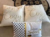 Three throw pillows on carpet: two large white pillows with gold script 'Let's get Cozy', one smaller pillow with black hearts, gold stripe, and 'Home Sweet Home' text