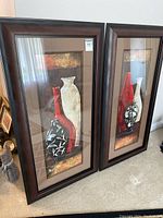 Two framed vertical wall art pieces leaning against a wall, each depicting three decorative vases in black, red and white with floral motifs.