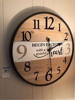 Front view of round wooden wall clock with black frame, large black numerals and inspirational quote in the center.