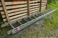 Full view of the 32-foot aluminum extension ladder laid on gravel next to wooden fencing.