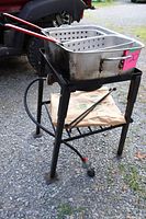 Side angle showing propane burner frame with oil pot and baskets on top, resting on gravel ground outside.