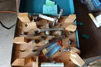 Photo showing a teal plastic case with assorted Makita router bits organized with wooden holders, a metal wrench, and other small parts inside. The case latch is broken, and the bits show signs of use.
