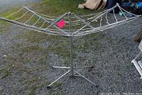 Photo of an old portable aluminium clothes line with four arms and cords on a tripod base outside on gravel ground.