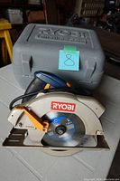 Photo of Ryobi circular saw and gray plastic carrying case on table. Saw blade visible with orange lever and power cord coiled.