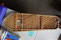 Full view of one wooden snowshoe showing the frame, woven rawhide webbing, and wooden handle.