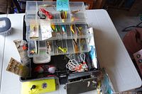 Overview of black tackle box open showing numerous compartments filled with assorted fishing lures and accessories, some still in packaging and others loose