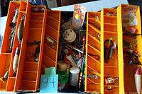 Top view of the open orange and black tackle box showing multiple compartments filled with various fishing lures, hooks, and other fishing accessories.