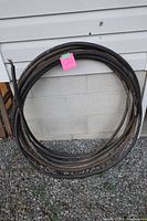 Coiled black armored cable labeled with pink tag '121' leaning against a cinder block wall outdoors on gravel ground.