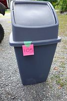 Blue plastic trash can with a revolving lid shown outside on gravel ground, labeled with a pink tag '105'.