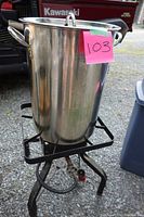 Tall metal frying pot with side handles and built-in thermometer, sitting on propane burner stand outdoors on gravel.