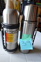 Two stainless steel vacuum flasks with carrying handles and caps, one with label ARCOSTE visible.