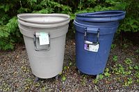Two Roughneck garbage cans side by side outdoors on gravel. One is beige and the other blue. Both have wheels and handles.