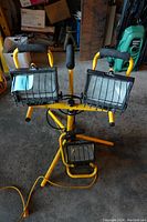 Full view of double halogen work light with adjustable tripod base and single 500 Watt light positioned below, yellow frames with black cage guards and foam handle grips.