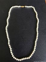 Full view of the necklace laid flat on a black surface, showing the entire single strand of white pearls and the gold clasp.