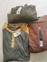Three pieces of plus size clothing including brown hoodie, olive green hoodie, and long sleeve yellow-grey henley shirt with tags visible laid flat.