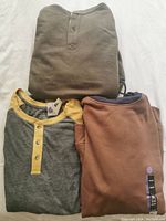 Three folded long sleeve shirts: grey with yellow accents, brown, and olive green.
