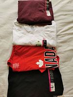 Four folded shirts shown: maroon, white V-neck, red with Canada print, maroon long sleeve with tags visible