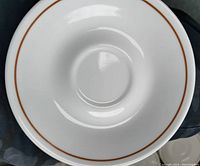 Close-up photo of a single white ceramic saucer with thin brown rim showing smooth glazed surface and cup indent.