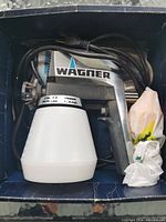 Overview of Wagner W320 paint spray gun in original box with power cord coiled and white spray container attached.