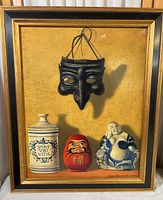 Framed print showing a black mask above three figurines (white jar, red Daruma doll, blue and white Buddha) against yellow background