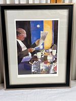 Full framed print of older man reading newspaper at a table with coffee cups and bowls. Signature visible in lower right. Frame in black with silver trim and white mat.