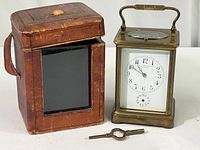 Clock in brass casing with white dial showing black numbers and smaller subsidiary dial, placed next to leather case and winding key.